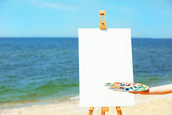 Female hand holding palette with paints and easel with canvas on beach — Stock Photo, Image