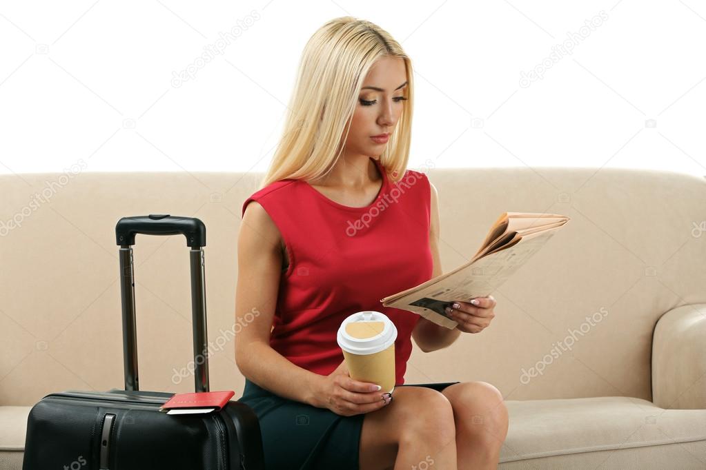Woman with suitcase and tablet sitting on sofa and reading newspaper isolated on white