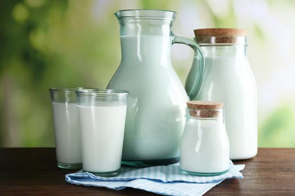 Dzban, słoiki i szklanki mleka na drewnianym stole, na tle przyrody — Zdjęcie stockowe