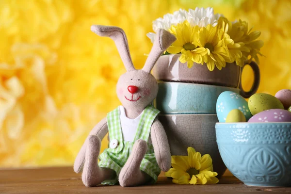 Пасхальный кролик с раскрашенными пасхальными яйцами с цветами на деревянном столе на желтом фоне — стоковое фото