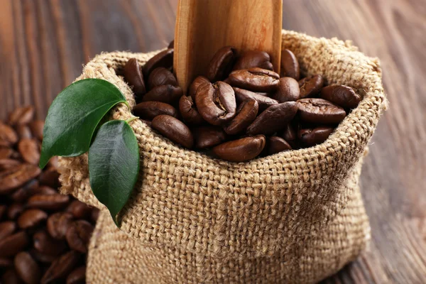 Grãos de café com folhas e colher no saco na mesa de madeira fechar — Fotografia de Stock