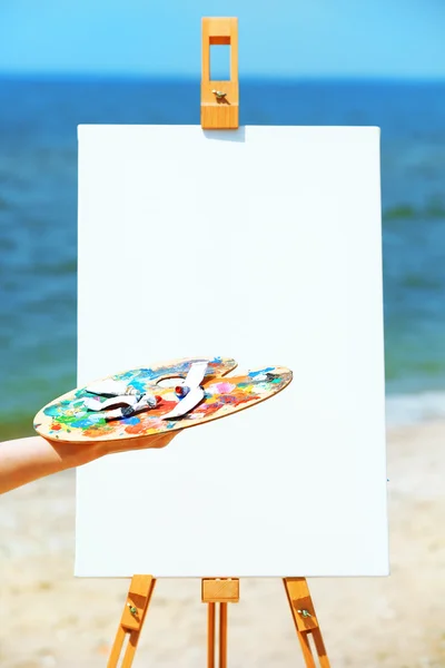 Female hand holding palette with paints and easel with canvas on beach — Stock Photo, Image