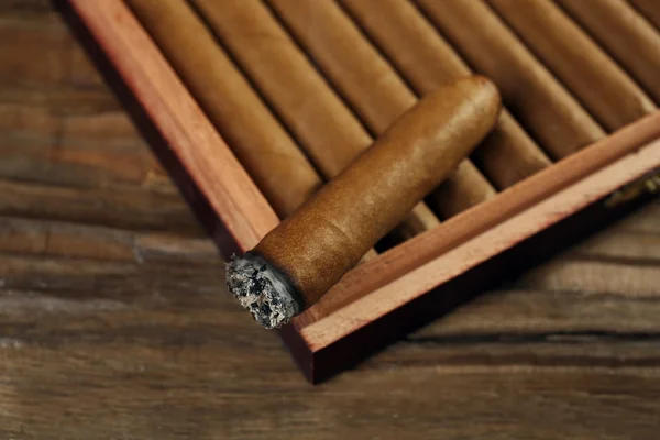 Cigars and burning one in box on wooden table, closeup — Stock Photo, Image