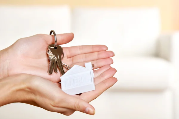 Female hands holding keys with house key chain on blurred sofa background — Stock Photo, Image