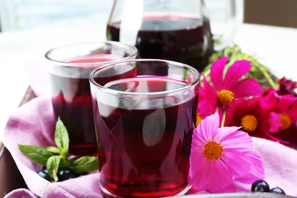 Glasses of fresh blackcurrant juice on wooden tray with pink napkin and flowers, closeup — Stock Photo, Image