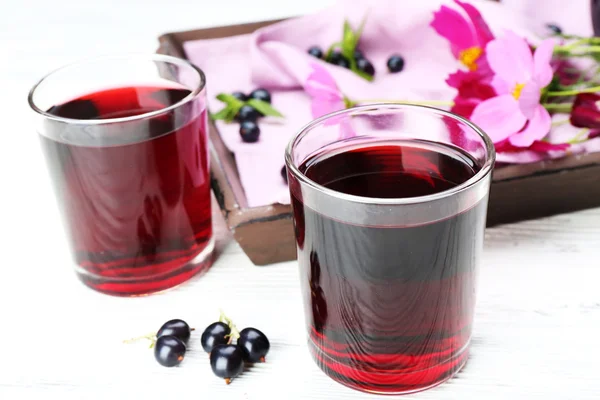 Glasses of fresh blackcurrant juice on wooden table near tray with flowers, closeup — Stock Photo, Image