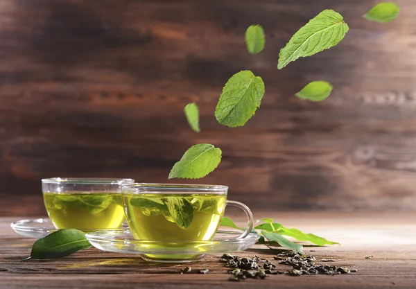 Mint leaves falling in cups of green tea on wooden background — Stock Photo, Image