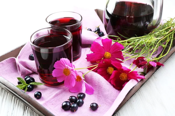 Стаканы свежего сока черной смородины на деревянном подносе с розовой салфеткой и цветами, крупным планом — стоковое фото