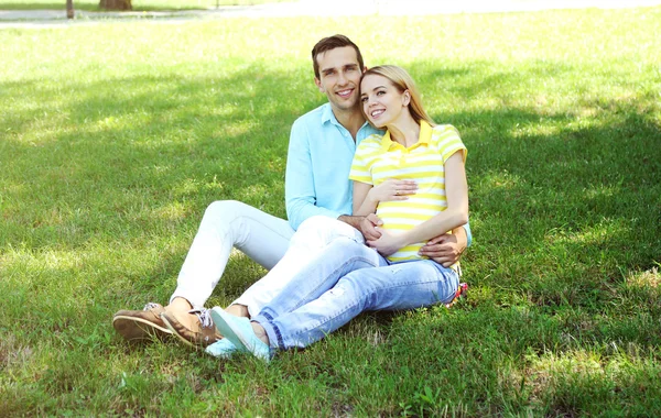 Молодая беременная женщина с мужем сидит на зеленой траве в парке — стоковое фото
