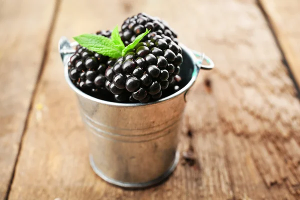 Ripe blackberries with green leaves in small metal bucket on wooden background