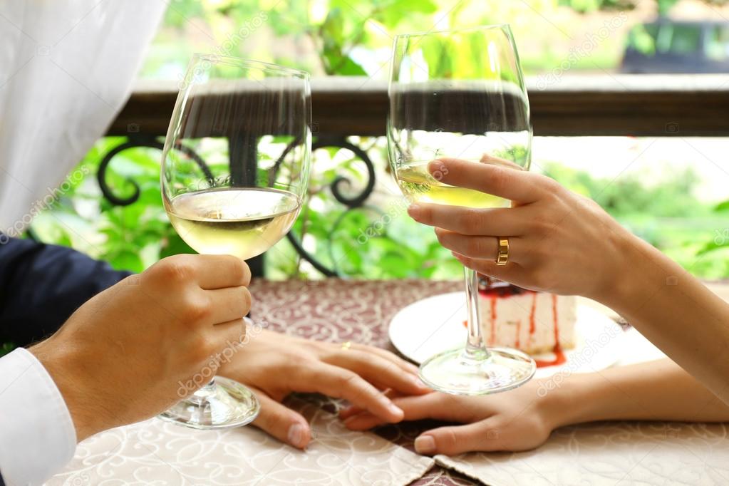 Hands of newlyweds holding glasses of wine at table in cafe, closeup