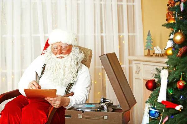 Santa claus siedzi z listy życzeń dzieci w wygodnym fotelu w pobliżu kominek w domu — Zdjęcie stockowe