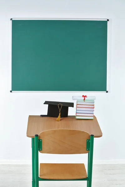 School desk with master hat, stack of books and diploma in classroom