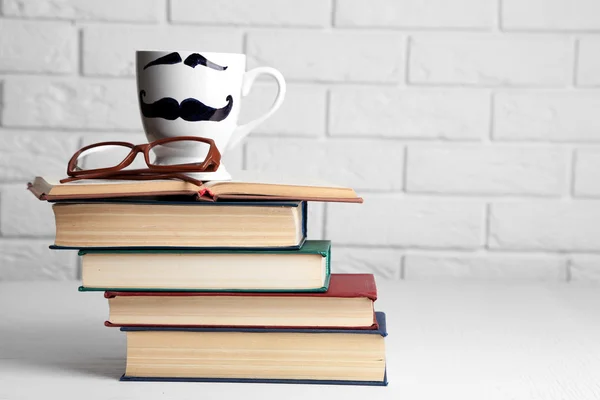 Винтажные книги и чашка с усами на деревянном столе на фоне кирпичной стены — стоковое фото