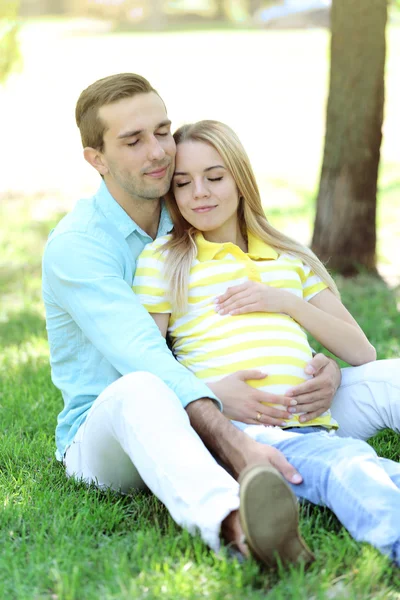 Молодая беременная женщина с мужем сидит на зеленой траве в парке — стоковое фото