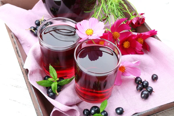 Glasses of fresh blackcurrant juice on wooden tray with pink napkin and flowers, closeup — Stock Photo, Image