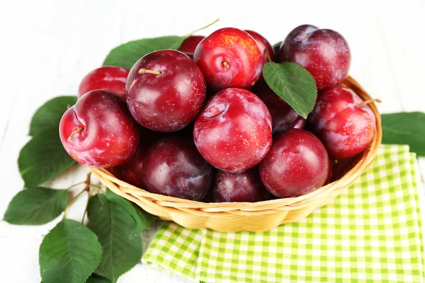 Ripe plums in wicker bowl on wooden table with napkin, closeup — Stock Photo, Image