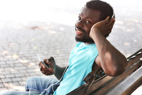 Hombre afroamericano escuchando música con auriculares en el banco en el parque —  Fotos de Stock