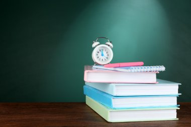 Masanın üzerinde yeşil kara tahta arka plan üzerinde kalem ve çalar saat ile kitap yığını