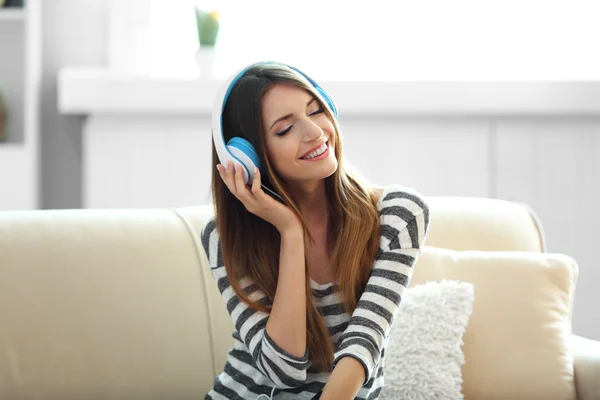 Mulher ouvindo música em fones de ouvido enquanto se senta no sofá no quarto — Fotografia de Stock