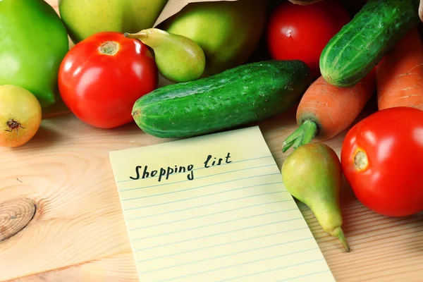 Heap of fruits and vegetables with shopping list on table close up