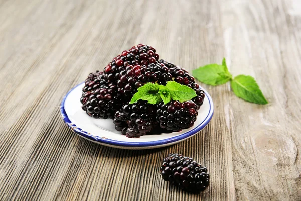 Ripe blackberries with green leaves in saucer on wooden background — ストック写真