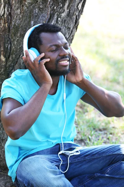 African American man listening music with headphones near tree in park — Stock Photo, Image