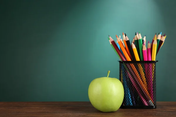 Soporte de metal con lápices de colores y manzana verde en el escritorio sobre fondo de pizarra verde —  Fotos de Stock