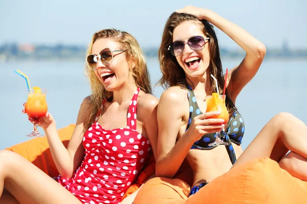 Two young girls enjoying cocktail on bag seats on beach at summertime — Stock Photo, Image