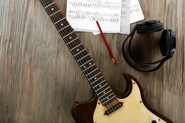 Electric guitar with headphones and music notes on wooden table close up — Stock Photo, Image