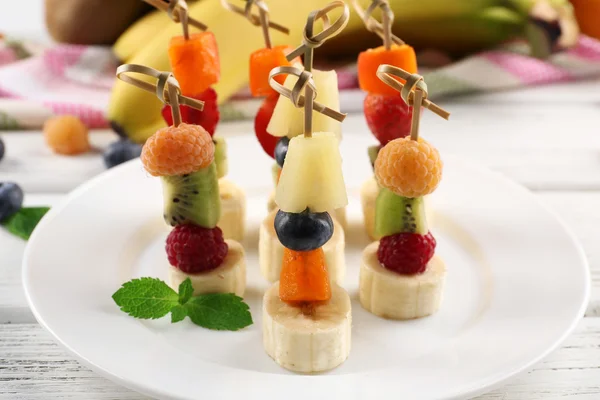 Fresh fruits on skewers in plate on wooden table, closeup