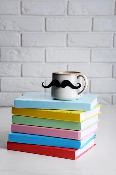 Bücher und Tasse mit Schnurrbart auf Holztisch auf Backsteinwand Hintergrund — Stockfoto