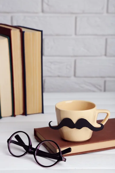 Vintage books and cup with mustache on wooden table on brick wall background — Stock Photo, Image