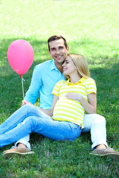 Mujer embarazada joven con marido sentado en la hierba verde en el parque —  Fotos de Stock