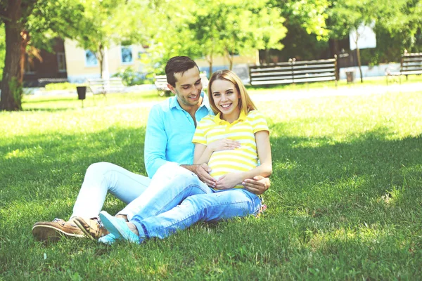 Mujer embarazada joven con marido sentado en la hierba verde en el parque —  Fotos de Stock