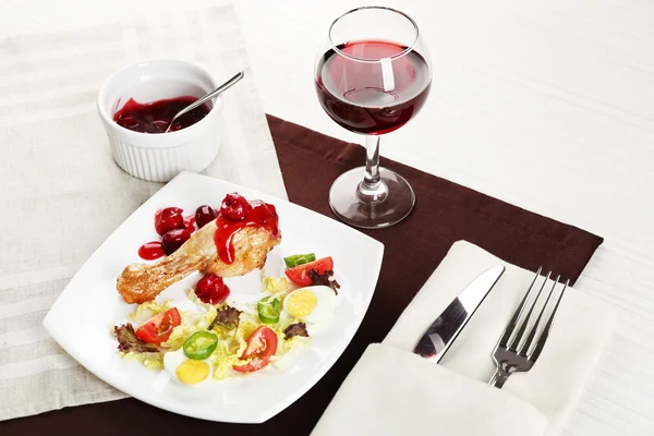 Dish of baked chicken leg and vegetable salad in white plate with glass of wine on table with napkin, closeup — Stock Photo, Image