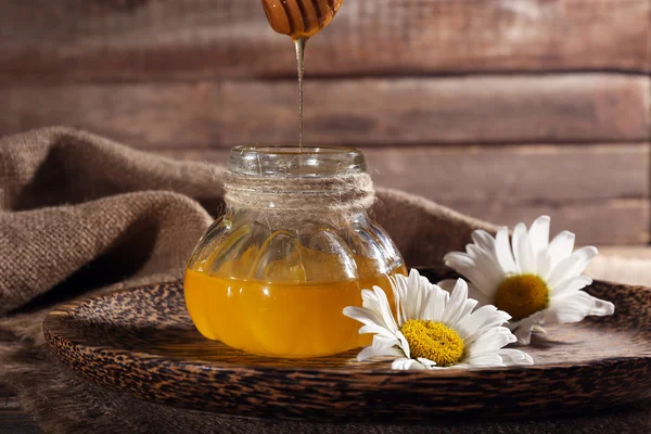 Miele versato da tuffatore di legno in vaso su vassoio con camomilla su sacco su fondo di legno — Foto Stock