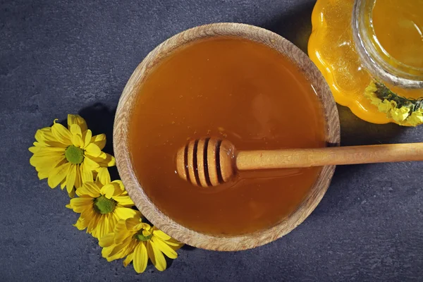 Honey in jar and in bowl, wooden dipper and flowers on dark grey background — Stock Photo, Image