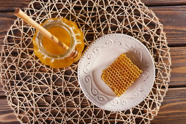 Maceta de miel con cazo de madera en ella y panal en placa sobre estera sobre fondo de madera — Foto de Stock