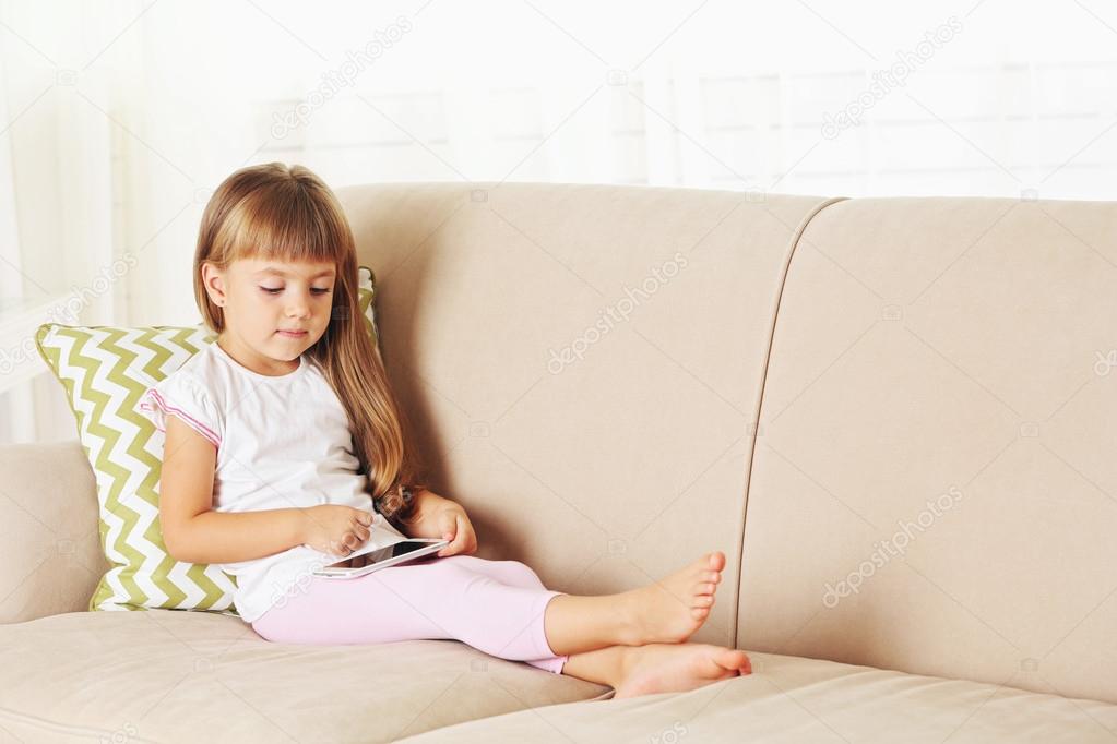 Beautiful little girl with smart phone sitting on sofa on home