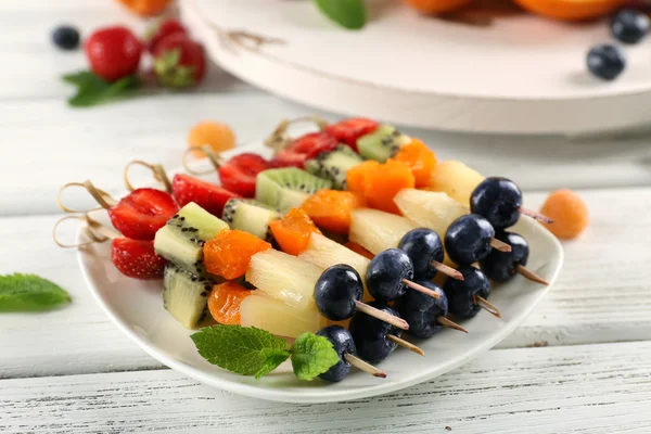 Fresh fruits on skewers in plate on wooden table, closeup — Stock Photo, Image