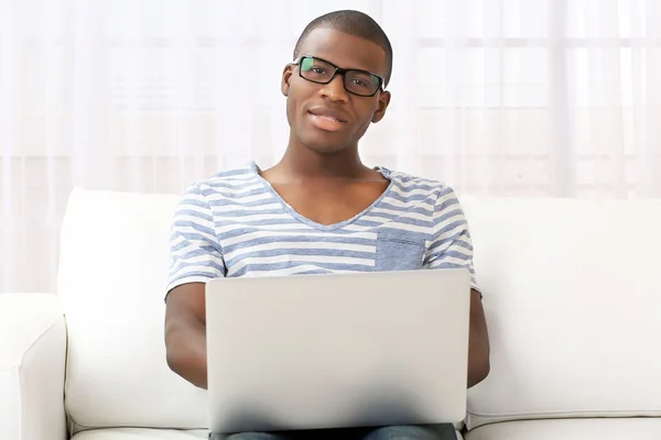 Hombre afroamericano guapo sentado con portátil en el sofá en la habitación —  Fotos de Stock