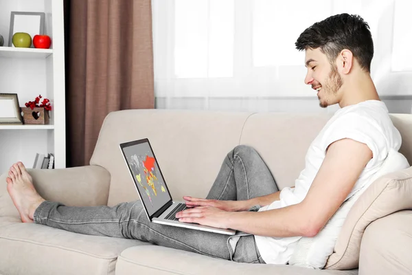 Handsome young man sitting on sofa and using laptop in room — Stock Photo, Image