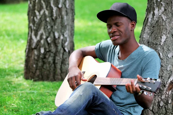 En kjekk afroamerikansk mann med gitar sittende i parken – stockfoto