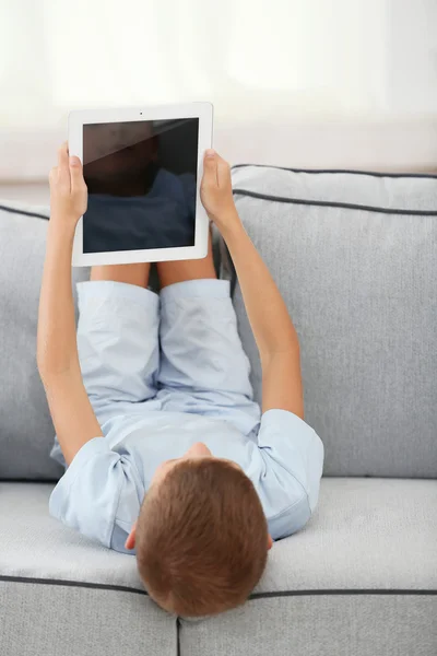 Dijital tabletli küçük çocuk kanepede oturuyor, evin arka planında. — Stok fotoğraf