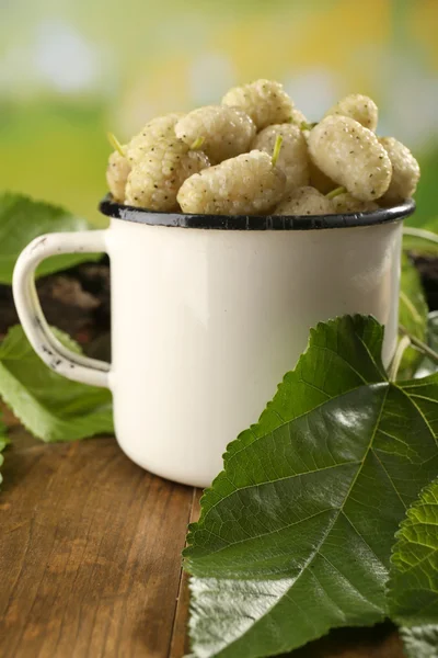 Ripe mulberries in mug with green leaves on table close up — Stock Photo, Image