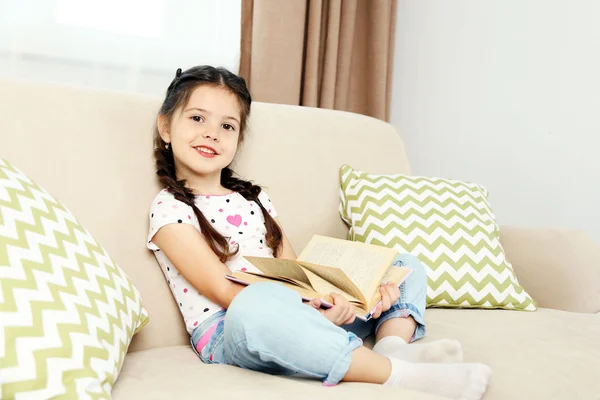 Красива маленька дівчинка сидить на дивані з книгою, на домашньому інтер'єрі фоні — стокове фото