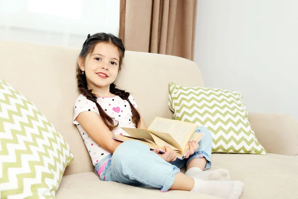 Vacker liten flicka sitter på soffan med boken, på hem inredning bakgrund — Stockfoto