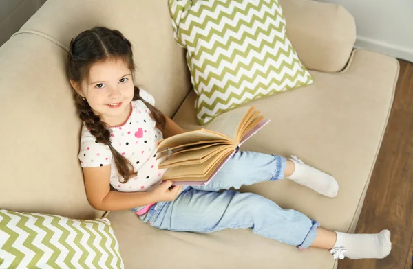 Vacker liten flicka sitter på soffan med boken, på hem inredning bakgrund — Stockfoto