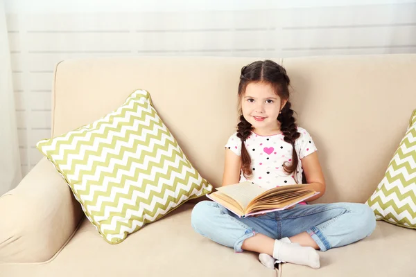 Hermosa niña sentada en el sofá con libro, en el fondo interior de casa —  Fotos de Stock